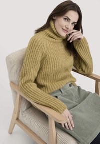 Frau mit braunen Haaren und blauen Augen sitzt in einem beigen Sessel, trägt einen senfgelben, grob gestrickten Rollkragenpullover und einen hellgrünen Cordrock.