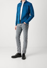Blue jacket with a high collar, black accents and zippers, paired with light gray jeans and black lace-up shoes. Simple, fitted design.