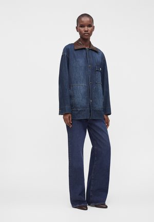 Personne debout portant une veste en jean bleu foncé avec un col marron et un pantalon en jean large assorti sur un fond clair uni.