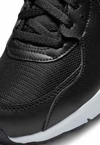 Parte superior de zapato deportivo negro que presenta un diseño de cuero sintético texturizado y malla, con cordones planos negros y una suela de goma blanca.