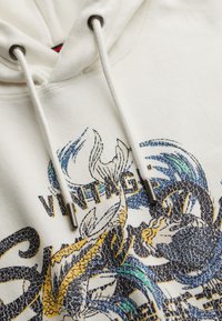 Sudadera color blanco roto con un diseño gráfico texturizado en azul, amarillo y negro. Incluye un cordón con aglets metálicos y un bolsillo tipo canguro.