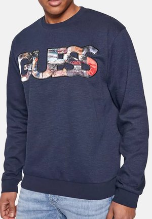 Man draagt een marineblauwe sweatshirt met een groot meerkleurig "GUESS" logo over de borst, gecombineerd met lichtblauwe jeans.