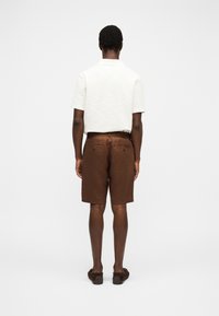 Homme debout de dos portant une chemise blanche à manches courtes, un short marron jusqu'aux genoux et des chaussures marron sans lacets.