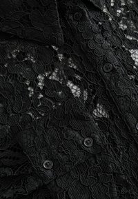 Black lace fabric features intricate floral patterns with a mix of textures. The garment has black buttons and a layered design.