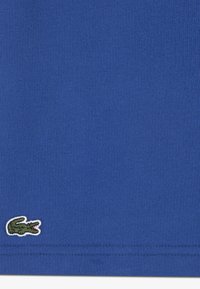 Sinise puuvillase t-särgi kangas, sileda tekstuuriga. Äärdele tikitud väike roheline krokodillilogo. Lihtne disain, ilma täiendavate mustriteta.