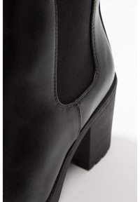 Mustat nahkasaappaat nilkkapituisella varrella ja paksulla korolla, joissa on joustavat sivupaneelit ja sileä pinta, korostaen ompeluyksityiskohtia.