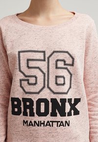 Felpa rosa con grandi numeri neri stampati "56" e "BRONX" con la parola "MANHATTAN" sotto. Realizzata in tessuto morbido con una finitura testurizzata.