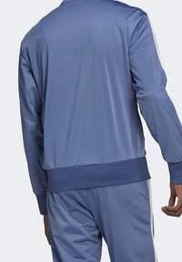 Vaaleansininen trikootakki, jossa on sileä pinta, resorivyötärö ja kontrastivalkoiset raidat hihoissa.