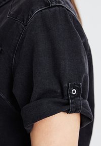 Zwarte denimshirt met opgerolde mouwen, voorzien van een knoopdetail. De stof is textuurrijk en stevig met een klassiek ontwerp.