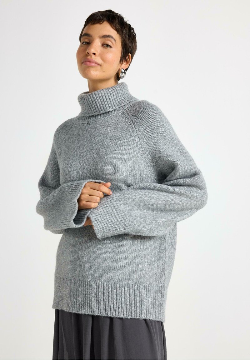 Person med kort hår iført en grå oversized rullekravesweater og mørk nederdel, der står foran en enkel hvid baggrund.