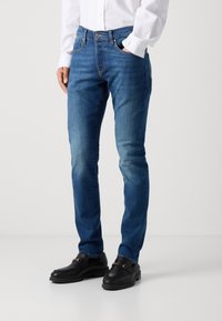 Blaue Denim-Jeans mit schlanker Passform, subtiler Ausbleichung, Fünf-Taschen-Design und Knopfverschluss. Getragen mit schwarzen Slippern.