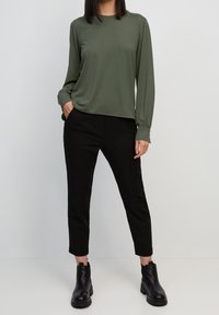 Maglietta verde oliva a maniche lunghe con polsi arricciati, abbinata a pantaloni neri slim-fit e stivaletti neri, su uno sfondo semplice.