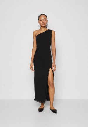 Robe noire asymétrique avec bordure en dentelle, fente haute sur la cuisse et coupe ajustée. Portée avec des chaussures plates noires, présentant un petit détail décoratif.