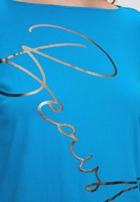 Niebieska bawełniana koszulka z krótkim rękawem, z luźnym krojem, która zapewnia komfort. Z przodu znajduje się abstrakcyjny design tekstu w metalicznie złotym kolorze.