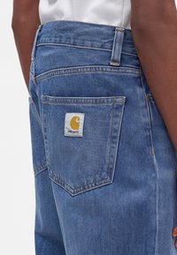 Τσέπη από μπλε τζιν με το λογότυπο Carhartt στην πλάτη, φορεμένη από άτομο με σκουρόχρωμο δέρμα και λευκό πουκάμισο.