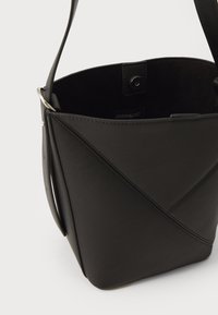 Melna ādas spaiņmaiss ar ģeometrisku šuvumu, regulējamu plecu siksnu, spiedpogu aizdari un gludu faktūru.