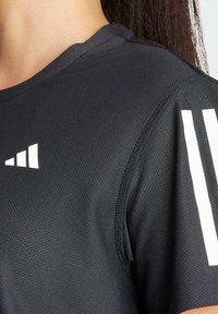 Svart sporttröja med textur, med vitt triangulärt märke på bröstet och två vita vertikala ränder på ärmen, buren av person med långt mörkt hår.