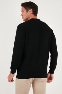 Melns ribots garajumsvārks ar noapaļotu kakla izgriezumu, ar sānu izgriezumiem un ribotiem piedurkņu galiem, redzams no aizmugures ar gaišiem biksēm.