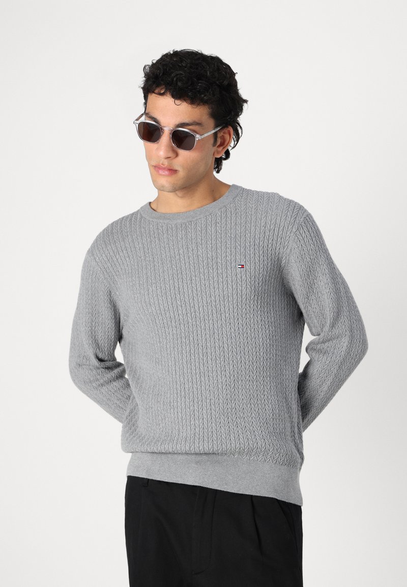 Pull en tricot gris avec un motif en grosse maille et un col rond. Présente un petit logo sur la poitrine. Porté avec des lunettes de soleil.
