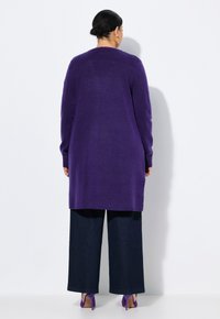 Lång lila kofta med mjuk textur, med långa ärmar och sidopartier, i kombination med vida mörka jeans och lila klackar.