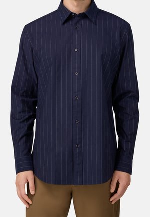 Homme portant une chemise bleu marine à manches longues, boutonnée, avec de fines rayures verticales blanches et un pantalon marron.
