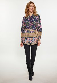 Blusa a fiori navy, con fiori verdi e rosa, dotata di una fascia dorata e una vita annodata; abbinata a pantaloni neri e stivali.