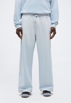 Mees, kes kannab helesiniseid laia säärega dressipükse, millel on nöörkinnitusega vöökoht, ja musti tossusid, seisab ühevärvilise tausta ees.