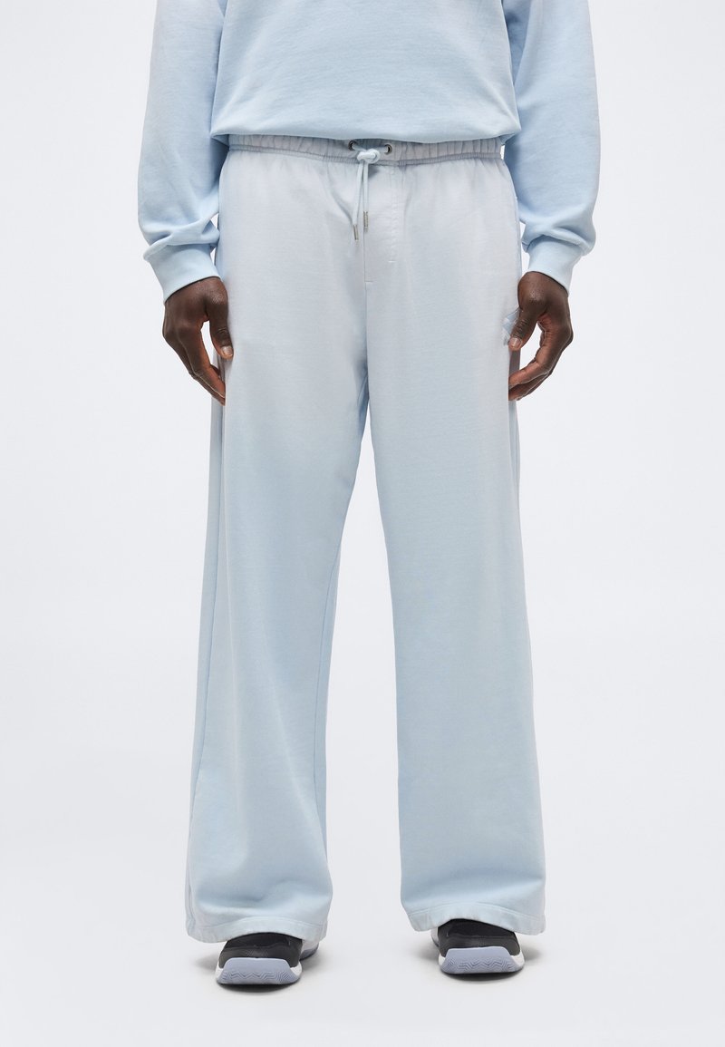 Άνδρας που φοράει ανοιχτό μπλε φαρδιά φόρμα με ρυθμιζόμενο κορδόνι στη μέση και μαύρα αθλητικά, στέκεται μπροστά σε απλό φόντο.