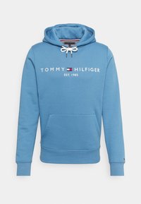 Modrá mikina s kapucí vyrobená z měkké tkaniny, s přední kapsou ve stylu klokan, stahovací kapucí a bílým nápisem "TOMMY HILFIGER EST. 1985" na hrudi.