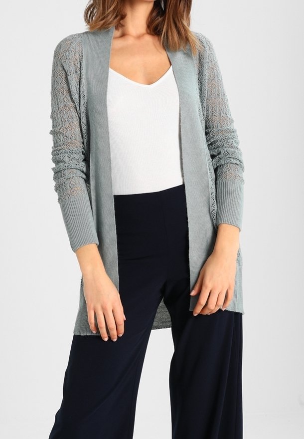 Cardigan en tricot bleu clair avec un motif en dentelle texturé sur les manches, design à ouvert à l'avant, porté par-dessus un haut blanc et un pantalon marine.