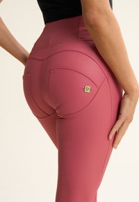 Leggings rosas de cintura alta hechos de material elástico. Presentan costuras curvas, dos bolsillos traseros y un pequeño logo metálico. Textura suave.