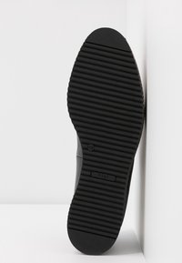 Semelle de chaussure en caoutchouc noir avec des rainures horizontales proéminentes et un bout arrondi. Présente un logo près du talon et un bord supérieur texturé.