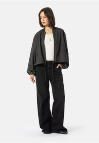 Sieviete ar plecu garuma tumšiem matiem, kas valkā brīvu melnu jaku, baltu topu, platmalu melnas bikses un zelta karoti.