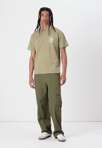 Olive green cotton t-shirt with a round logo on the chest, paired with matching cargo pants featuring side pockets and a relaxed fit.