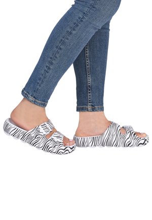 Pieds portant un jean bleu et des sandales blanches à enfiler avec des rayures zèbre noires et deux brides à boucle, vus de côté.