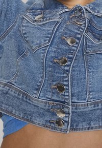 Detailní záběr modré džínové bundy s kovovými knoflíky označenými nápisem "ONLY" a pod ní modrý textilní oděv.