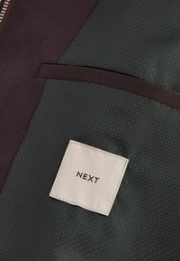 Jachetă dintr-un material verde închis și burgund, cu textură hexagonală, o pungă pe piept și un eticheta pătrată cu "NEXT" imprimat în negru.