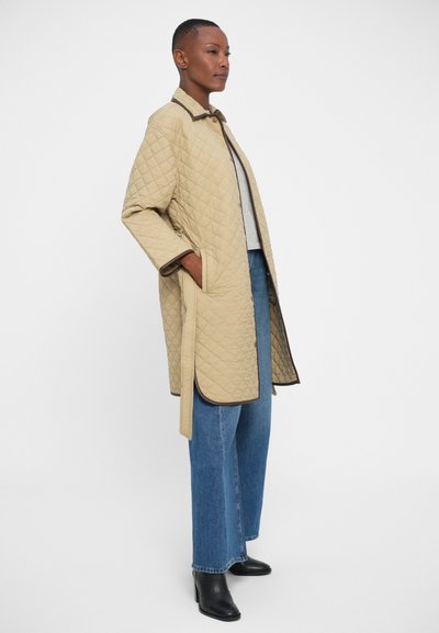 Beige quilted coat with brown trim, featuring side pockets, a button closure, and a relaxed fit, worn over a gray top and blue jeans.