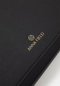 Gros plan sur une surface en cuir noir texturé avec un logo circulaire doré et le nom de la marque "ANNA FIELD" en lettres dorées.