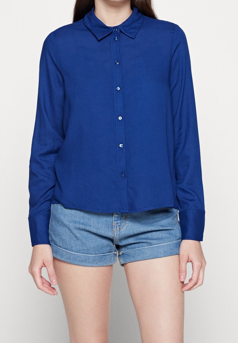 Chemise bleue à manches longues avec un col et une fermeture à boutons. Tissu doux, ourlet légèrement court, associée à un short en jean.