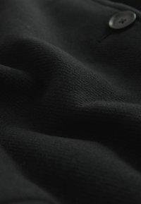 Tissu en laine noire avec une surface douce et texturée ; présente un petit bouton noir ; apparence légèrement froissée mettant en valeur son drapé.