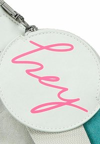 Etichetta rotonda bianca in pelle con testo in corsivo rosa "hey," attaccata a una cintura con tessuto teal e accenti in tessuto grigio. Hardware in argento visibile.