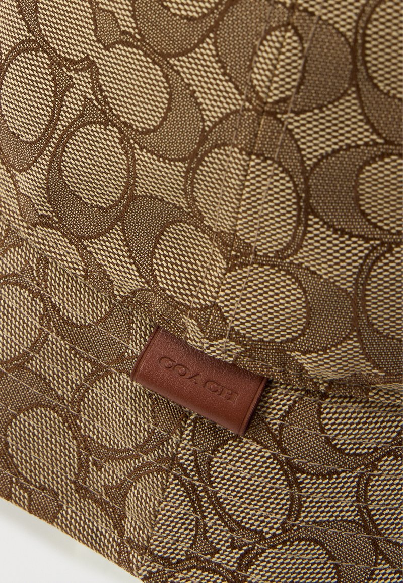 Brunt stof med et indviklet cirkelmønster og teksturerede accenter. Et syet lædermærkat viser mærkenavnet "COACH."