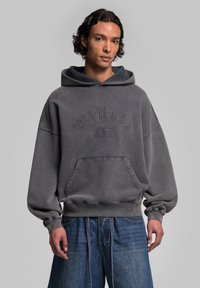 Sweatshirt à capuche surdimensionné gris avec une poche avant et un logo brodé. Il présente une texture douce et une coupe décontractée, stylisé avec un short en denim large.