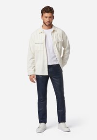 Lichtbeige corduroy shirt over een wit T-shirt, gecombineerd met donkerblauwe slim-fit jeans en witte sneakers. Het shirt heeft twee borstzakken.