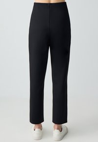 Pantalons noirs taille haute avec une coupe droite. Présente une texture lisse et un dos sans couture. Portés avec des baskets blanches.