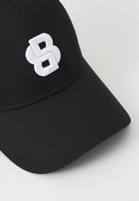 Fekete baseball sapka fehér hímzett "B" logóval. A dizájn tartalmaz egy strukturált koronát és ívelt napellenzőt varrási részletekkel.