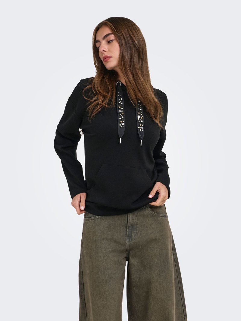 Femme portant un sweat à capuche noir avec des cordons décorés et un pantalon large vert olive, debout les mains près des poches sur un fond uni.