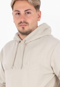 Felpa leggera beige realizzata in materiale morbido, con un logo ricamato e un design ad albero sul petto. Cappuccio con stringhe regolabili.