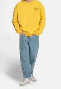 Yellow sweatshirt with a round neckline and front logo, paired with light blue loose-fitting jeans and white sneakers.